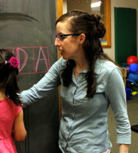 A woman stands at a chalkboard on which is written various letters. 