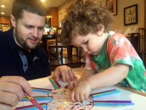 A toddler places small stickers onto a piece of paper as his dad watches on. He has a look of focused concentration on his face.