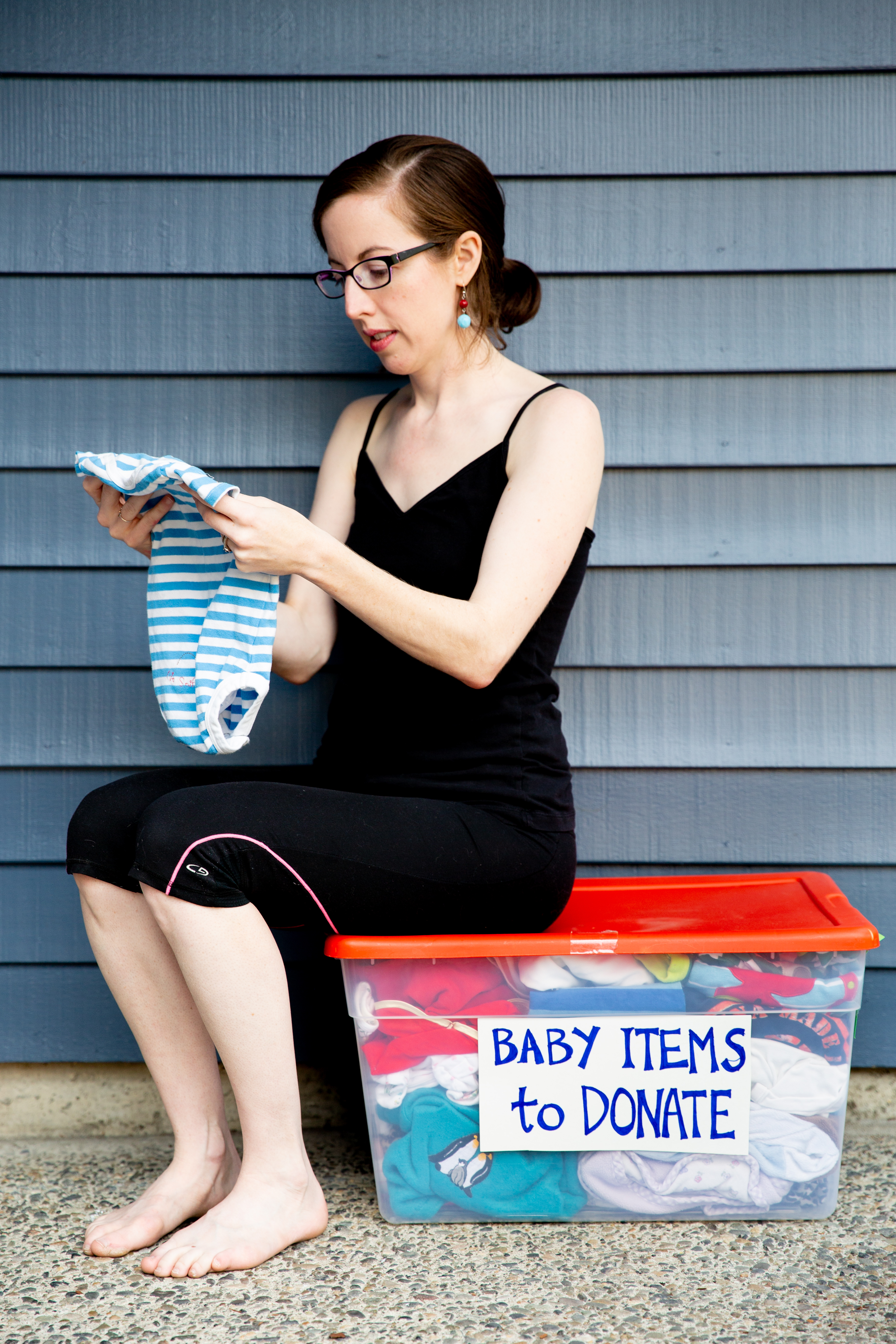 A woman sits on top of a box which has the label, "Baby clothes to donate." She looks at a small onesie.