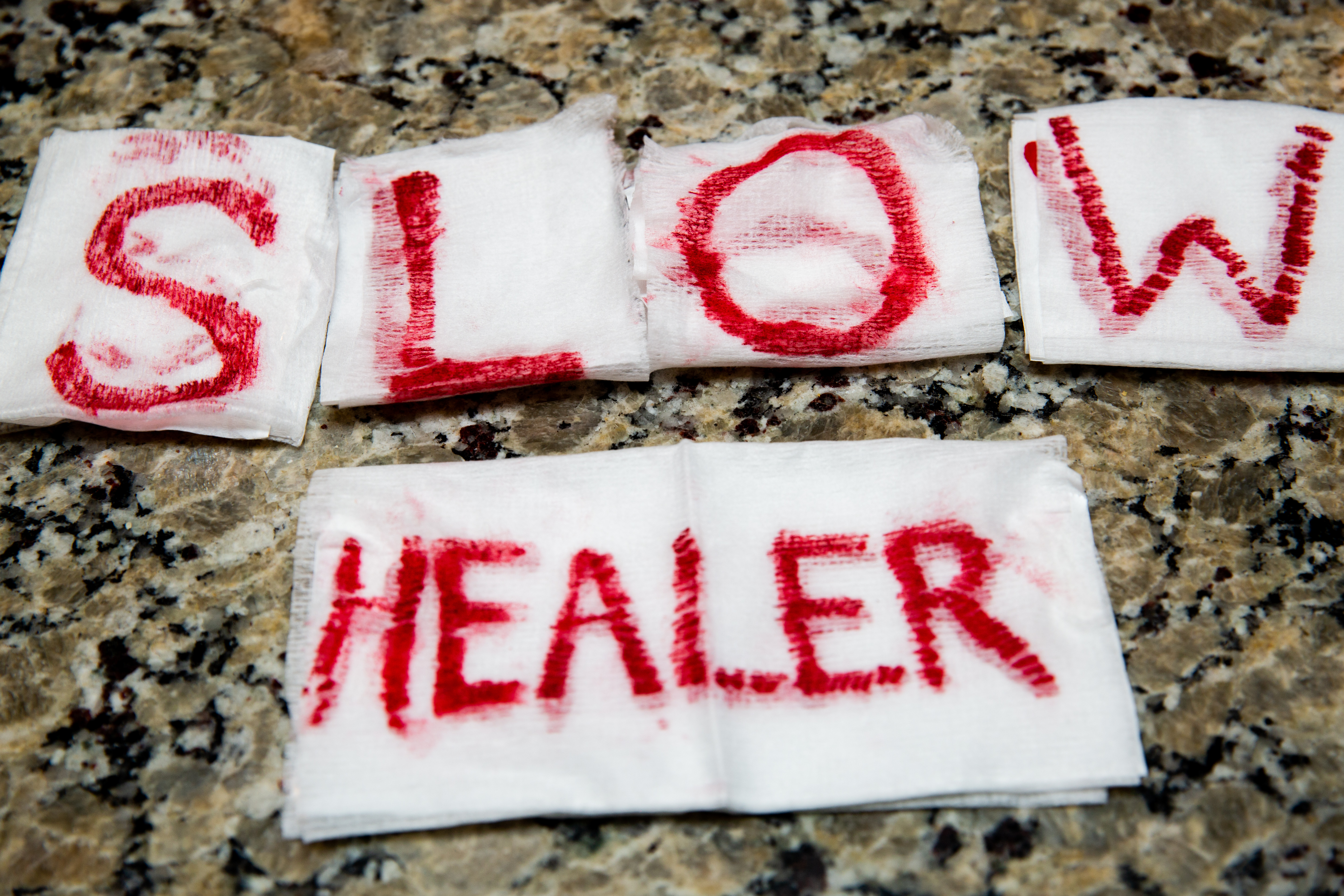 On 6 different white gauze bandages are written the letters for the words "slow healing," in what looks like blood. 